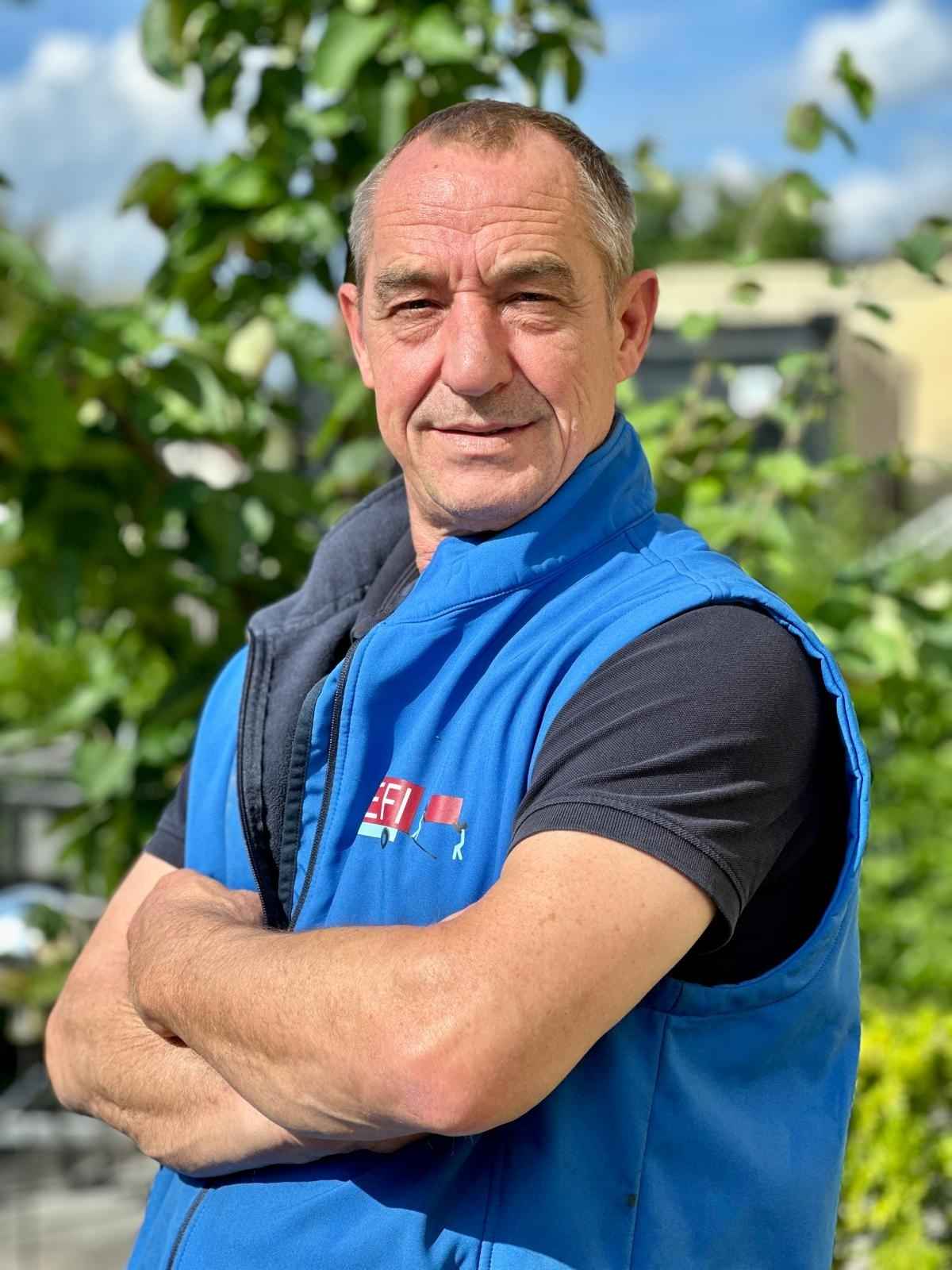 Mitarbeiter im blauen Arbeitsgilet mit verschränkten Armen im Grünen – Teamfoto von Efi Transporte in Würzburg.