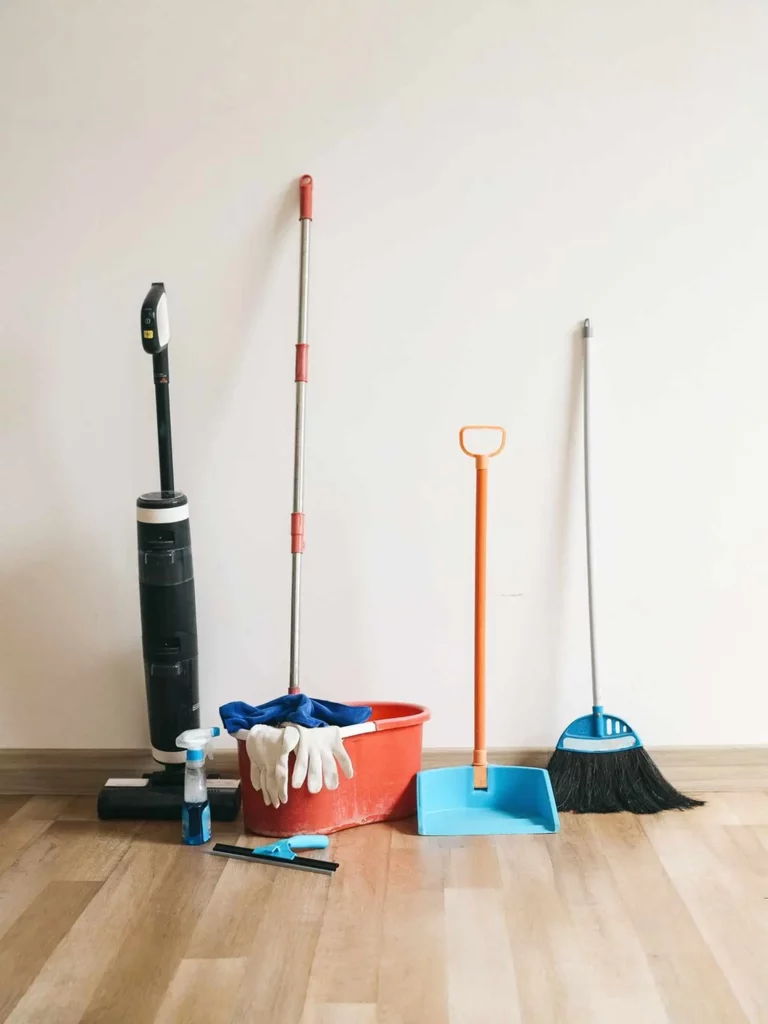 Reinigungsutensilien – Staubsauger, Eimer, Handschuhe, Kehrschaufel und Besen – stehen in einem leeren Raum; alles für die besenreine Übergabe bereit.