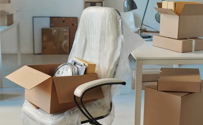 Büroauflösung: in Folie verpackter Bürostuhl, geöffneter Karton mit Wanduhr und Unterlagen, weitere Kartons am Schreibtisch – Räumung vor dem Abtransport.